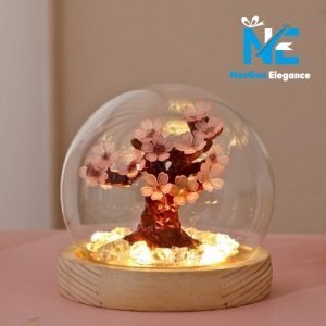 A close-up of a small, decorative cherry blossom tree inside a clear glass dome. The tree has light pink flowers on dark branches and is set on a light wood base. The base contains small, clear crystals that are lit from beneath, creating a warm glow. The logo "NexGen Elegance" is in the top right corner.