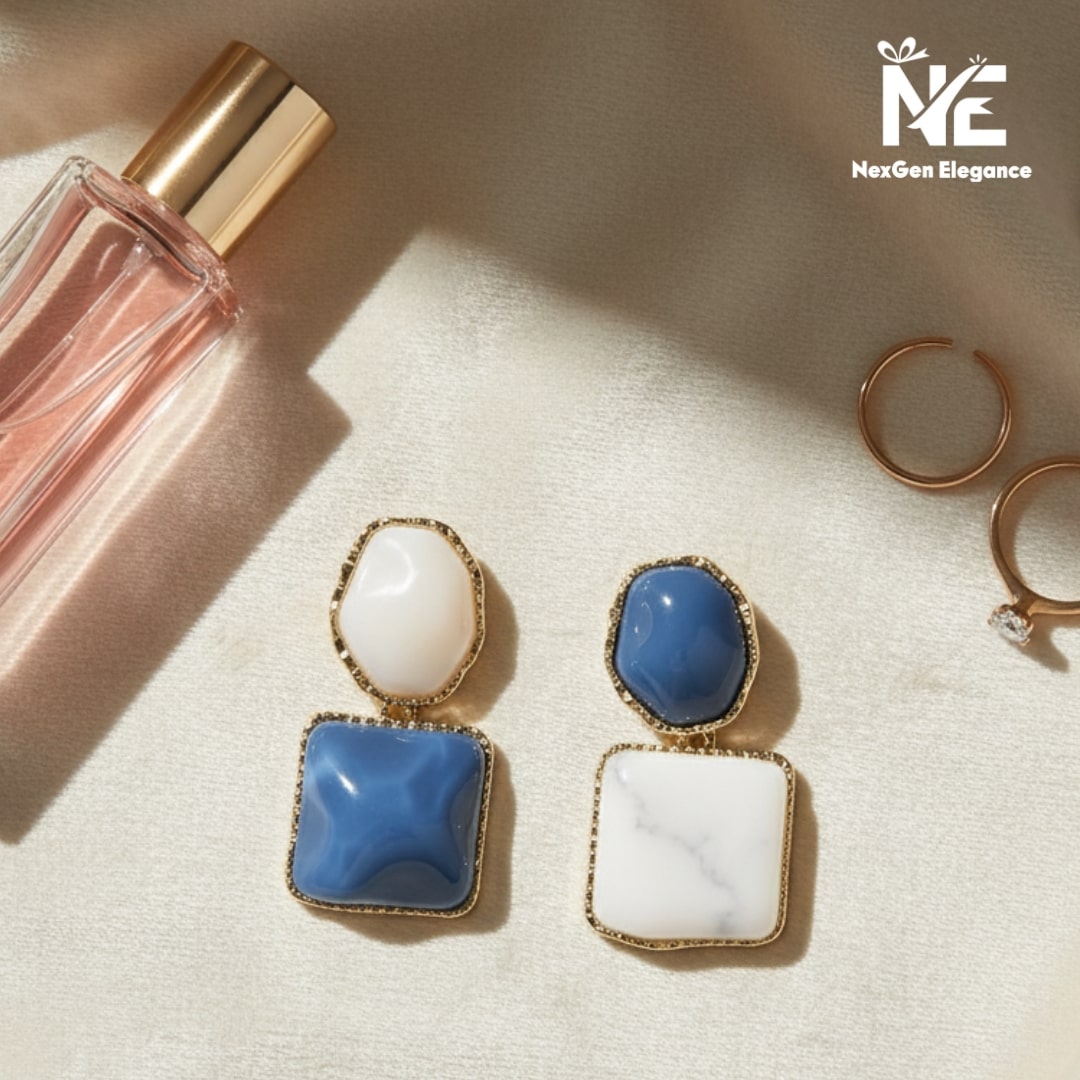 Top view of geometric blue and white earrings arranged on beige velvet cloth with perfume bottle and gold rings.